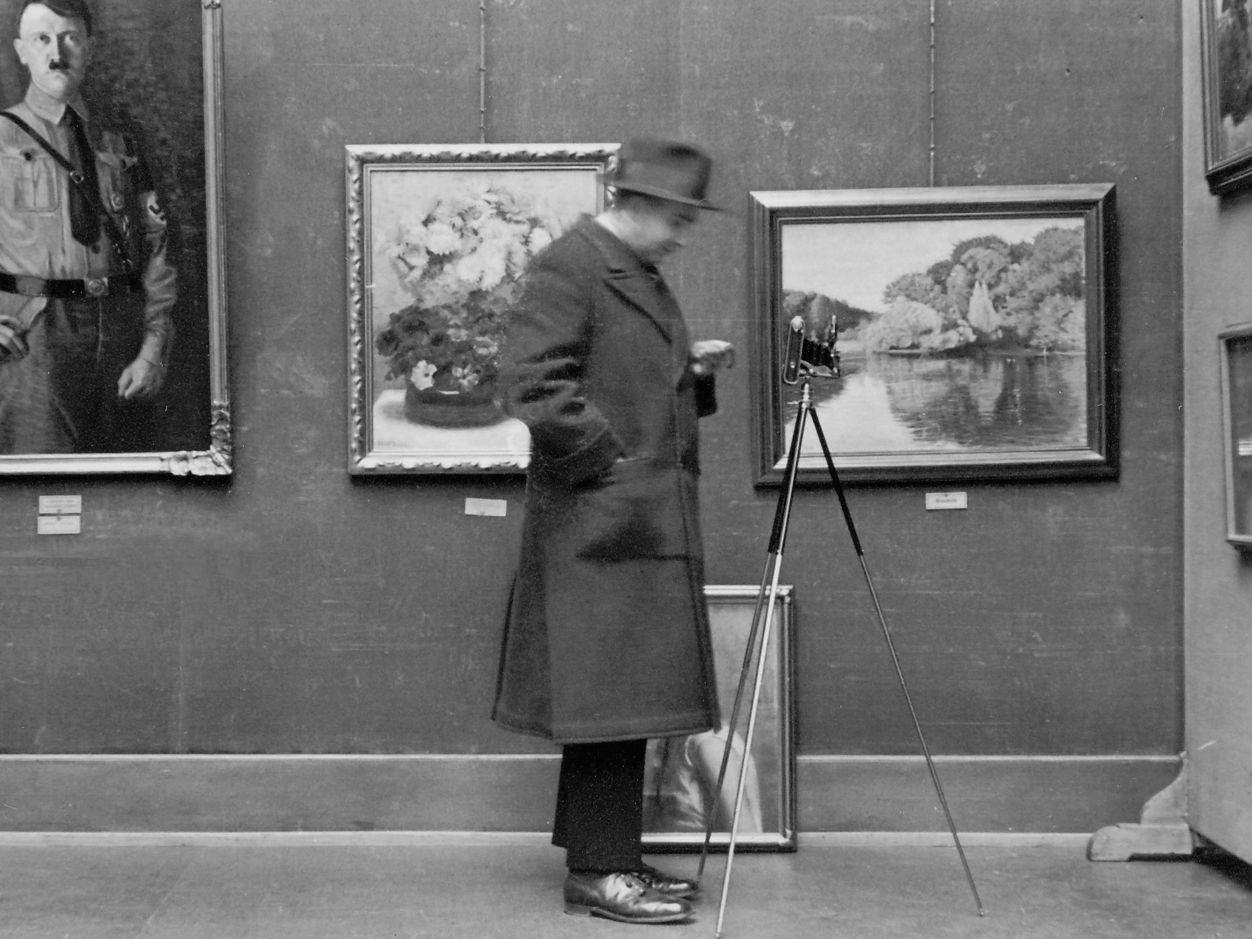 Schwarz-Weiß Fotografie eines Mannes mit Hut und Stativ vor einer Bilderwand (Im Hintergrund gerahmte Bilder)