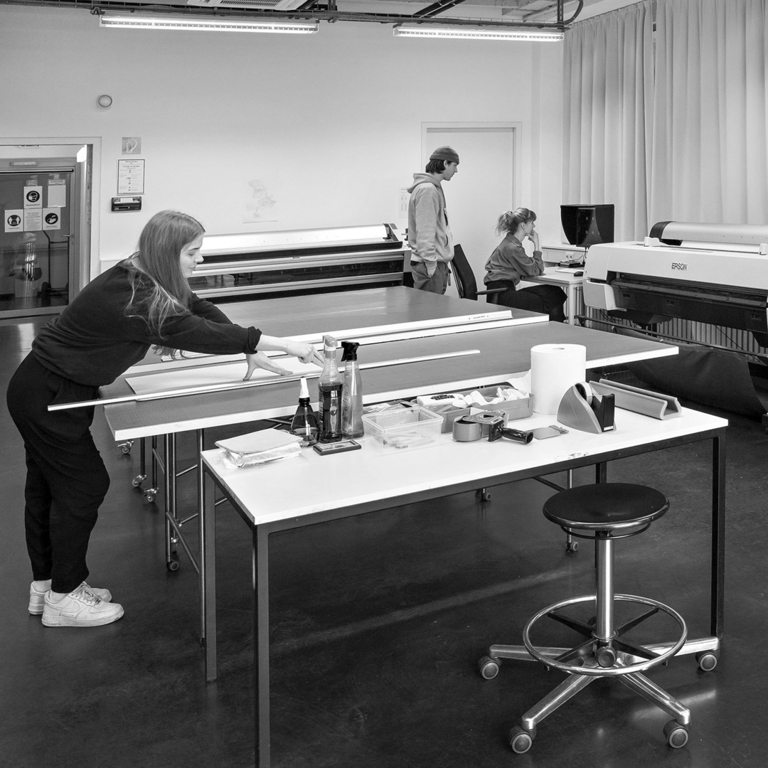 S/W Fotolabor: Eine Frau steht im Vordergrund und arbeitet an einem großen Tisch mit Lineal und Werkzeugen, während zwei weitere Personen im Hintergrund an einem Computer und einem Großformatdrucker beschäftigt sind.